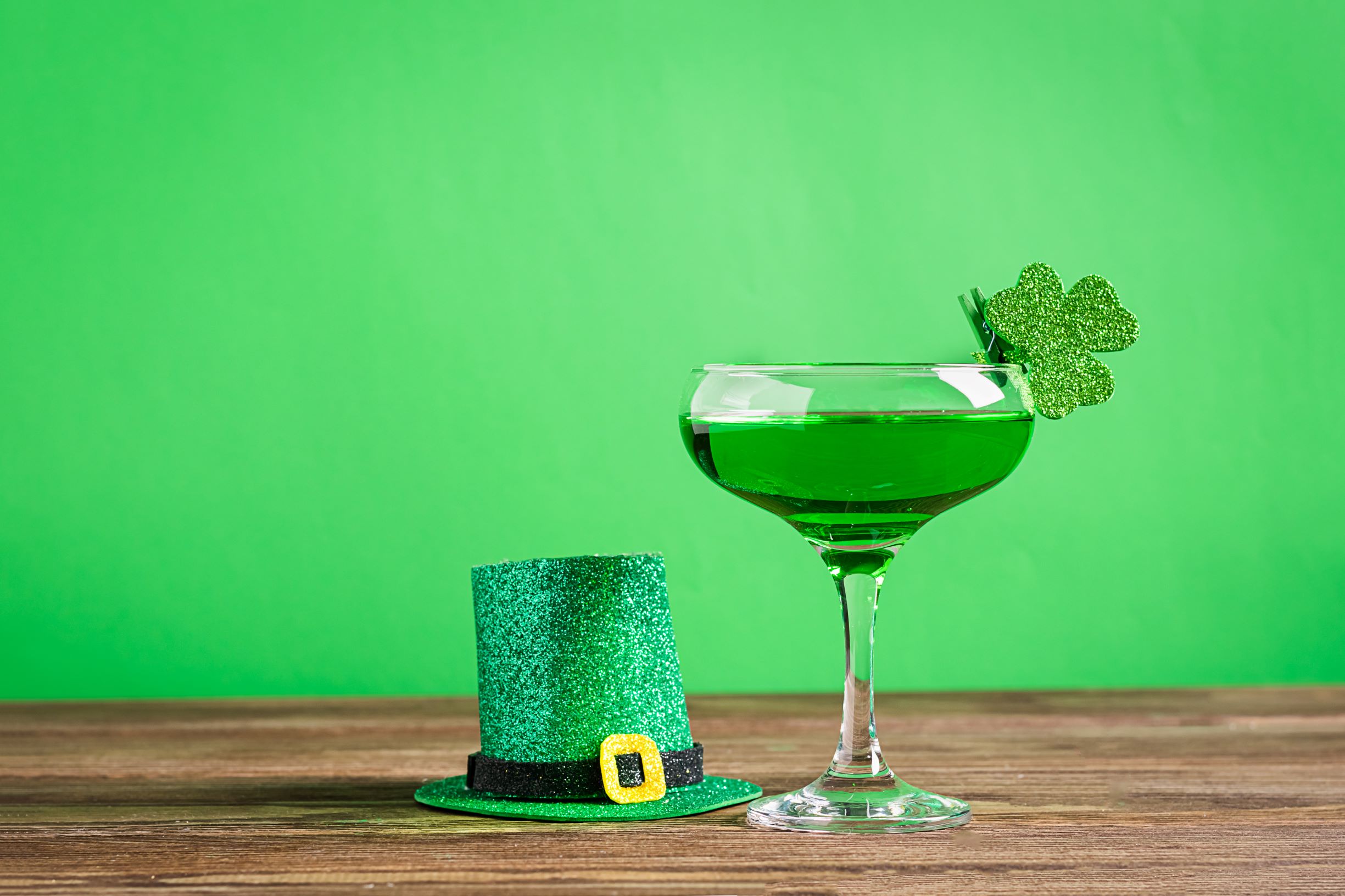 A green cocktail with a sparkly green clover clipped to the rim sits next to a small, sparkly leprechaun hat in front of a green background for St. Patrick's Day.